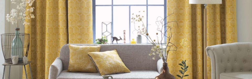 Bild Gardinen - Helles Wohnzimmer mit gelb gemusterten Vorhängen, einem grauen Sofa mit gelben Kissen und dekorativen Pflanzen neben einem großen Fenster. Firmenadresse