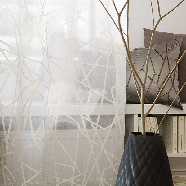 Transparente Gardinen mit geometrischem Muster in einem modernen Innenraumambiente, mit einer Vase mit Zweigen und einem Blick auf ein Bücherregal. Firmenadresse