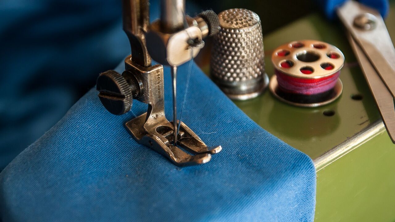 Gardinennäherei - Nähservice -Nahaufnahme der Nadel und des Nähfußes einer Nähmaschine auf blauem Stoff, mit Schere, Fingerhut und Garnrollen in der Nähe. Firmenadresse