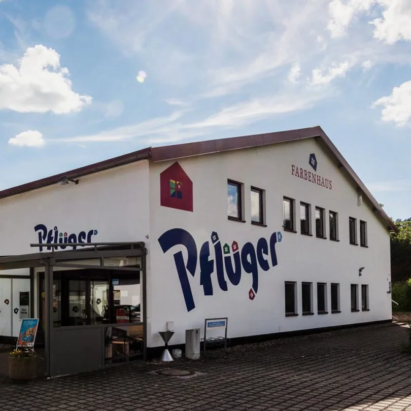 Ein weißes zweistöckiges Gebäude mit den Aufschriften „Pflüger“, „FARBENHAUS“ und „Schads Wohnträume“ an der Außenseite, mit vielen Fenstern und einer gepflasterten Fläche davor unter einem leicht bewölkten Himmel. - Firmenadresse