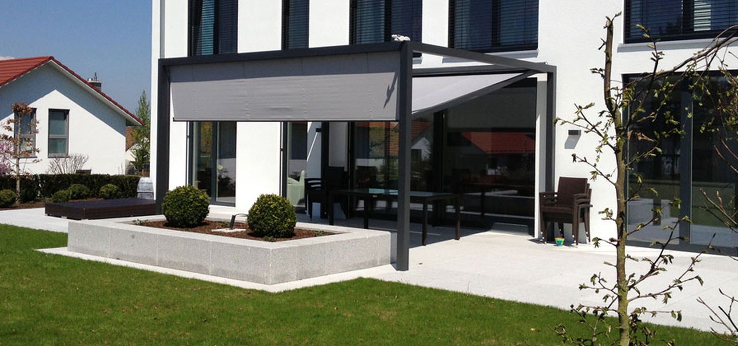Modernes weißes Haus mit großen Fenstern, einem überdachten Terrassenbereich mit Markisen und Sitzgelegenheiten im Freien, Hochbeeten und einem gepflegten Rasen. - Firmenadresse