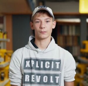 Fabio Koch mit einem weißen Kapuzenpullover mit der Aufschrift „XPLICIT REVOLT“ und einer weißen Mütze steht im Innenbereich, im Hintergrund sind Regale und Werkzeuge zu sehen, die den mutigen Geist von Über Uns widerspiegeln. - Firmenadresse