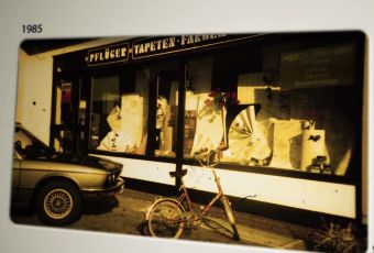 Ein Fahrrad steht auf dem Bürgersteig vor einem Geschäft mit großen Schaufenstern, in denen verschiedene Artikel ausgestellt sind. Links ist ein Auto teilweise zu sehen. Das Foto mit der Überschrift „1985“ zeigt eine klassische Straßenszene – perfekt für eine Einführung in „Über Uns“. - Firmenadresse