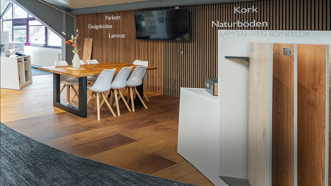 Moderner Ausstellungsraum mit einem Esstisch aus Holz und weißen Stühlen, Wänden aus Holzpaneelen, ausgestellten Bodenbelagsmustern und einem an der Wand montierten Fernseher – perfekt, um unsere Produkte und Serviceoptionen zu erkunden. - Firmenadresse