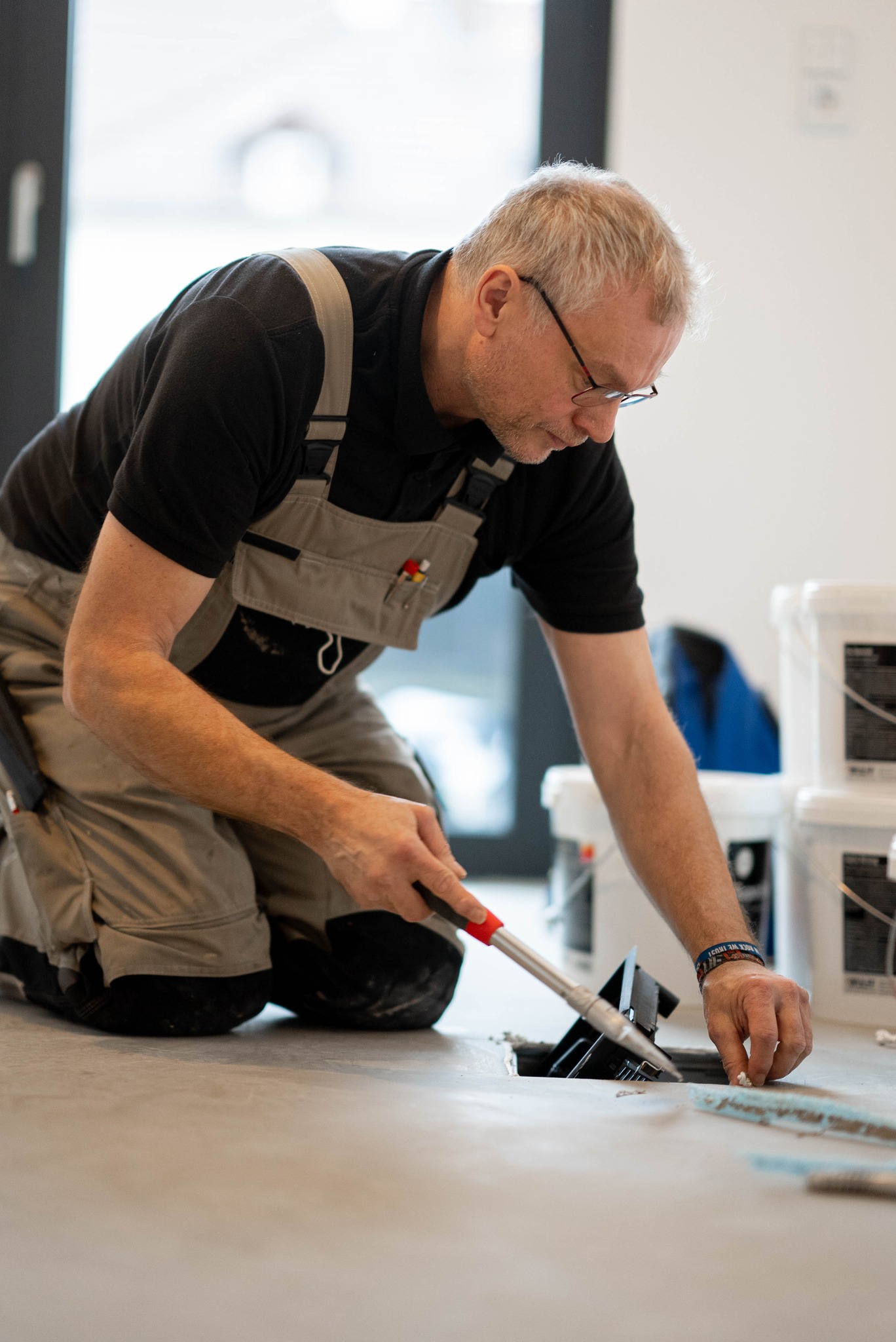 Ein Mann im Arbeitsoverall kniet auf dem Boden und trägt mit einer Kelle Klebstoff oder Fugenmasse auf, während er im Innenbereich Fußböden verlegt – ein wahrer Ausdruck unseres Engagements bei Über Uns für handwerkliches Können. - Firmenadresse