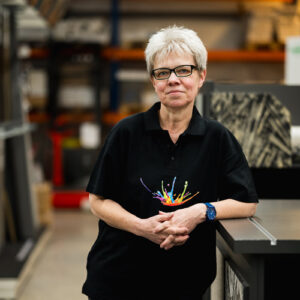 Eine Frau mit kurzen grauen Haaren und Brille steht in einem Arbeitsbereich. Sie trägt ein schwarzes Poloshirt mit einem farbenfrohen „Über Uns“-Design und eine blaue Uhr. Im Hintergrund sind Regale und Materialien zu sehen. - Firmenadresse