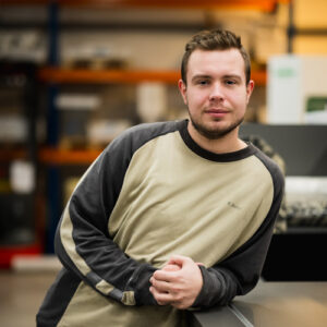 Ein Mann mit kurzen Haaren und gestutztem Bart, der ein beige-graues Langarmhemd trägt, lehnt in einem Industrieumfeld an einer Theke und spiegelt den fleißigen Geist hinter unserer Über Uns-Geschichte wider. - Firmenadresse