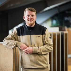 Kevin Hainke mit kurzen blonden Haaren und Bart steht drinnen, lehnt an einem Holzdisplay und trägt einen hellbraunen und schwarzen Pullover mit der Aufschrift „K. Halilaj“, und fängt mit seiner einladenden Präsenz den Geist von Über Uns ein. - Firmenadresse