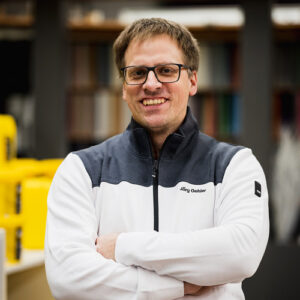 Jörg Oehler mit Brille und einer weiß-grauen Jacke mit der Aufschrift „Jörg Oehler“ steht mit verschränkten Armen im Haus, lächelt in die Kamera und ist bereit, seine Geschichte für die Rubrik „Über uns“ zu erzählen. - Firmenadresse