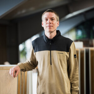 Dennis Knierim in einem beige-schwarzen Pullover steht drinnen, stützt eine Hand auf eine Holzoberfläche und blickt direkt in die Kamera – und fängt den zugänglichen Geist hinter unserer Über-Uns-Geschichte ein. - Firmenadresse
