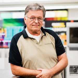 Ein älterer Mann mit grauem Haar und Brille, der ein beige-schwarzes Poloshirt trägt, steht im Innenbereich vor einem unscharfen, bunten Hintergrund – perfekt für eine Über-Uns-Einführung. - Firmenadresse