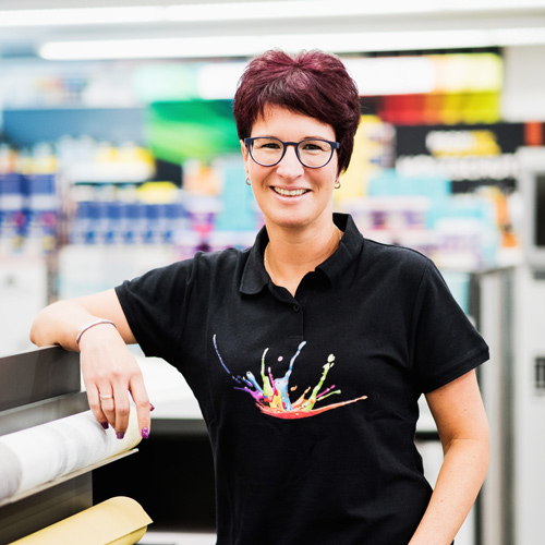 Melanie Kenn mit kurzen dunklen Haaren und Brille steht drinnen. Sie trägt ein schwarzes Hemd mit einem bunten Spritzermuster und steht neben einer großen Papierrolle – perfekt für den Abschnitt „Über uns“. - Firmenadresse