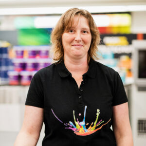 Eine Frau in einem schwarzen Poloshirt mit buntem Spritzerdesign steht in einem Ladengang mit Farbdosen im Hintergrund und verkörpert den kreativen Geist unserer Über Uns-Geschichte. - Firmenadresse