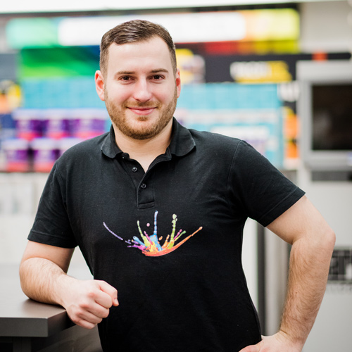 Ein Mann mit kurzen Haaren und Bart steht im Haus und lächelt. Er trägt ein schwarzes Poloshirt mit einem bunten Spritzer-Motiv. Einen Arm stützt er auf die Theke und verkörpert den freundlichen Geist unseres Über Uns-Teams. - Firmenadresse