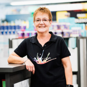 Eine Frau mit kurzen Haaren und Brille steht lächelnd an einer Theke im „Über Uns“-Bereich des Ladens und trägt ein schwarzes Poloshirt mit einem bunten Farbspritzer-Design. - Firmenadresse
