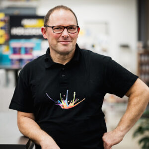 Ein Mann mit kurzen Haaren und Brille trägt ein schwarzes Poloshirt mit buntem Spritzerdesign, steht drinnen, einen Arm auf einer Theke abgestützt, und verkörpert den freundlichen und zugänglichen Geist unseres Über Uns-Teams. - Firmenadresse