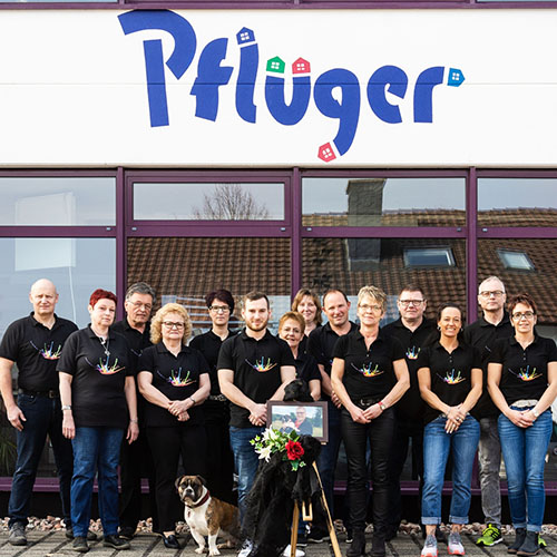 Team Farbenhaus Pflüger in passenden schwarzen Hemden steht vor einem Gebäude mit der Aufschrift "Pfluger", mit zwei Hunden und einem gerahmten Foto mit Blumen in der Mitte, versammelt für eine besondere Kontaktveranstaltung. - Firmenadresse